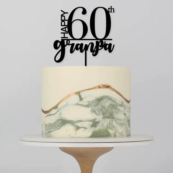 A round cake decorated with a smooth white icing. The bottom half features a marbled design with shades of green and a gold stripe. On top of the cake, there is a black acrylic cake topper that reads "HAPPY 60th grandpa." The cake is placed on a simple white cake stand. The background is a plain light color.