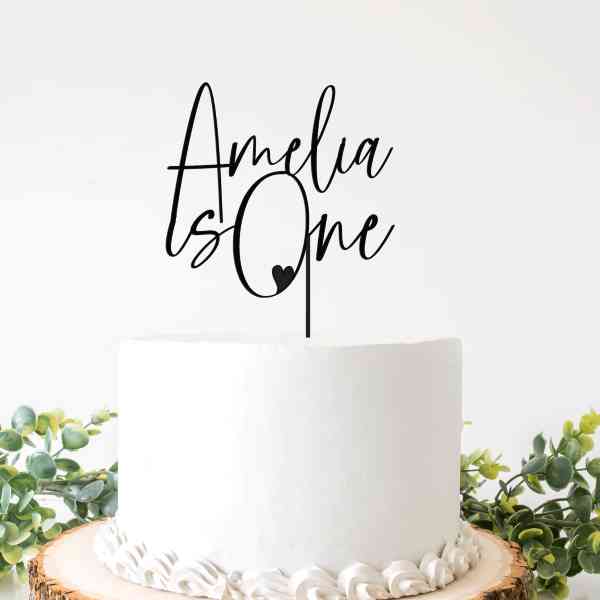 A white round cake topped with a black acrylic cake topper that reads "Amelia is One" in stylized font. The cake is decorated with white frosting around the edges, and is placed on a wooden slice with greenery around it. The background is light and neutral, enhancing the cake&