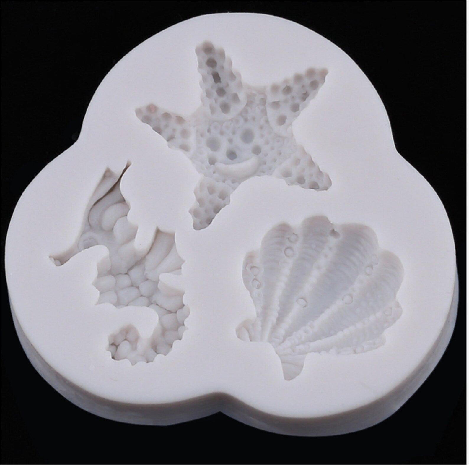 The image shows a white silicone mold designed for creating decorative shapes. The mold features three cavities in the shapes of a seahorse, a starfish, and a shell. Each design has detailed textures, representing realistic features of each marine creature. The mold is rounded and has a smooth finish, suitable for baking or crafting purposes. The background is black, highlighting the white silicone material of the mold.
