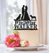A custom black acrylic cake topper featuring a silhouette of a couple standing together, with two dogs next to them. The text "Mr. & Mrs. Walker" is prominently displayed below the couple. The topper is placed on a white cake adorned with pink peonies and smaller flowers, resting on a decorative plate against a softly blurred background.