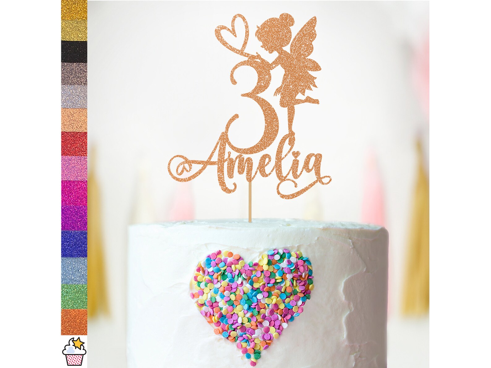 A decorative cake topper featuring a glittery fairy figure beside the number "3" and the name "Amelia," displayed on a white frosted cake. The cake is adorned with colorful heart-shaped sprinkles in shades of pink, blue, and yellow. To the left, a color palette of glitter cardstock options is shown in various vibrant hues, including gold, silver, black, and several shades of pink and purple. The background features soft, blurred decorations.
