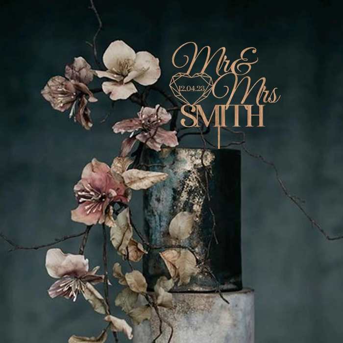 A decorative acrylic cake topper featuring the words "Mr & Mrs" with the last name "SMITH" and the date "12.04.23" is displayed. The topper is adorned with a diamond graphic and is set against a background of muted colors. It is integrated with a floral arrangement of soft pink and white flowers, along with twisting branches, adding a natural and elegant touch to the presentation. The topper is placed atop a textured, cylindrical cake stand.