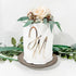 A decorated white cake topped with artificial flowers, including a light-colored rose and greenery. The cake features a shiny gold acrylic charm with the initials "JM" on its side. The cake sits on a round brown cake stand, surrounded by greenery. The background is a simple light color.