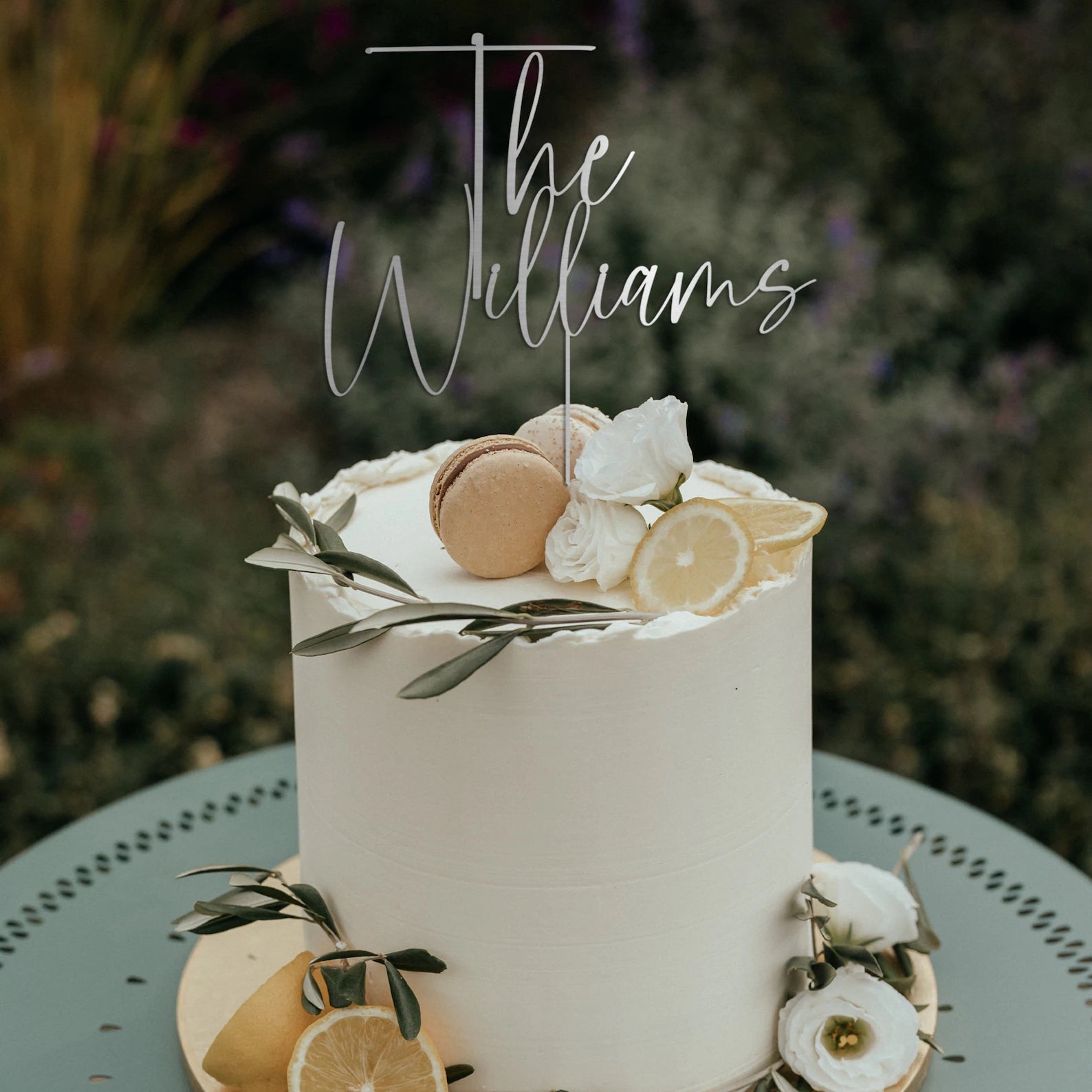 A decorated white wedding cake featuring a customized acrylic topper that reads "The Williams." The cake is adorned with macarons, slices of lemon, and white roses along the top and sides, with green leaves artistically arranged around it. The cake is displayed on a decorative table amidst a floral background.