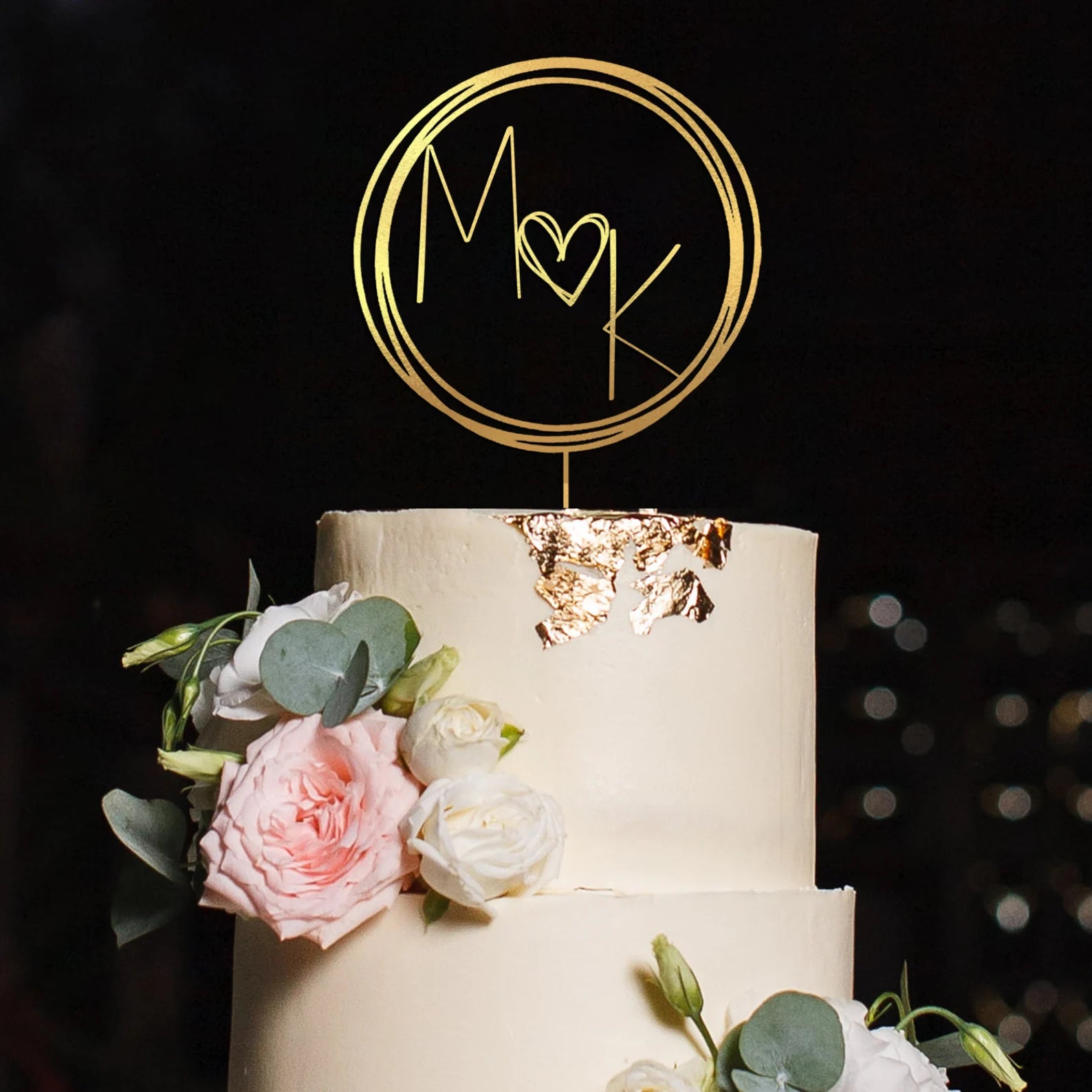 A three-tiered white wedding cake topped with a gold acrylic cake topper featuring the initials "M" and "K" inside a circular design with a heart in between. The cake is adorned with delicate pink and white flowers, as well as eucalyptus leaves, and has gold leaf accents on the top tier. The background is dark, highlighting the cake and its decorations.