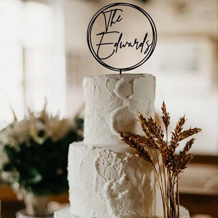 A three-tiered white wedding cake adorned with textured icing. Atop the cake is a black acrylic cake topper featuring the name "The Edwards" inside a circular design. Surrounding the base of the cake are dried wheat-like plants, set against a softly lit background that suggests an elegant event setting.
