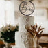 A three-tiered white wedding cake adorned with textured icing. Atop the cake is a black acrylic cake topper featuring the name "The Edwards" inside a circular design. Surrounding the base of the cake are dried wheat-like plants, set against a softly lit background that suggests an elegant event setting.