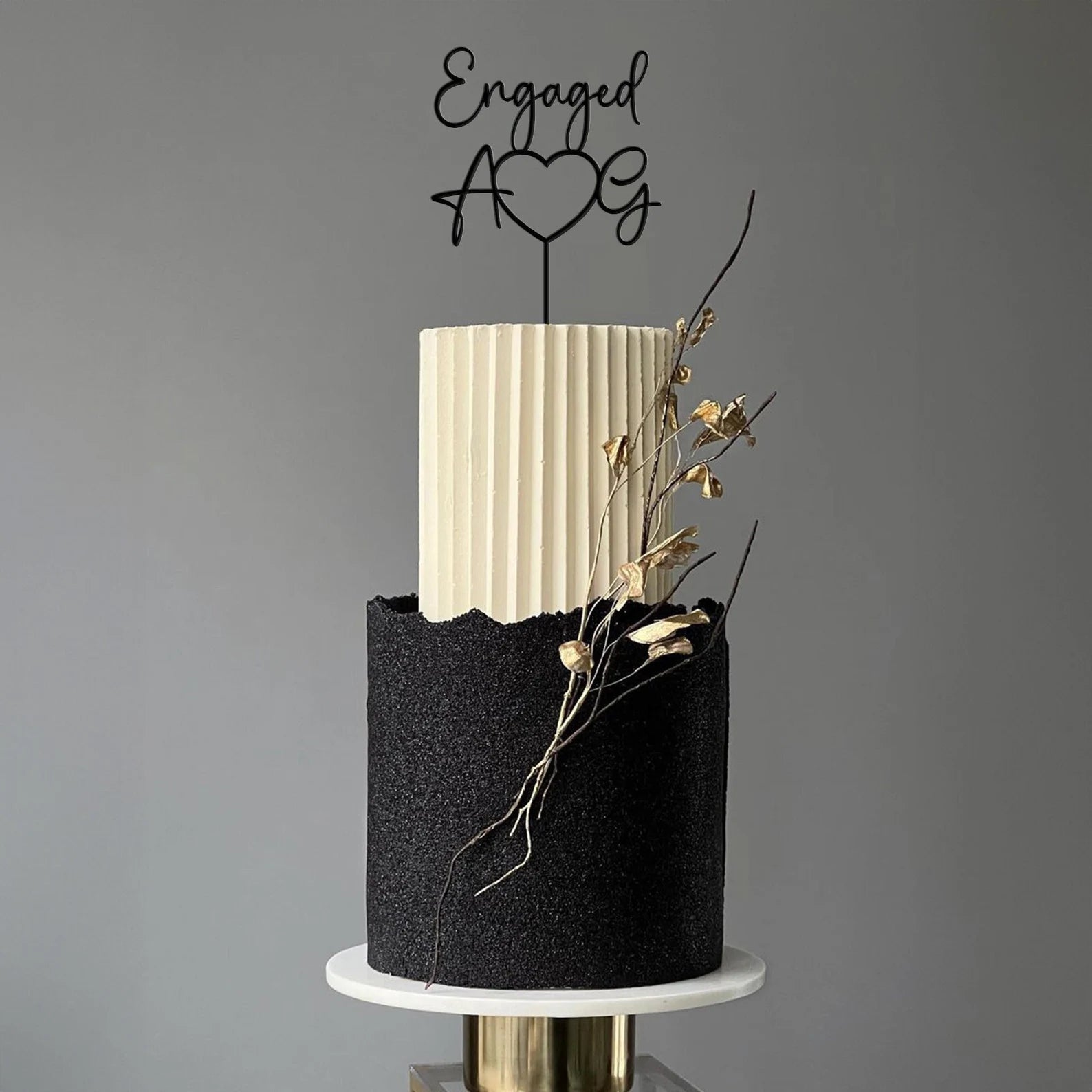 A modern engagement cake featuring a two-tier design. The top tier is a tall, ribbed white layer, while the bottom tier is a textured black layer with a wavy edge. Atop the cake is a stylish acrylic topper reading "Engaged" inside a heart shape with the letters "A" and "G" positioned next to it. The cake is adorned with slender, gold-accented twigs and a minimalist base, set against a neutral background.