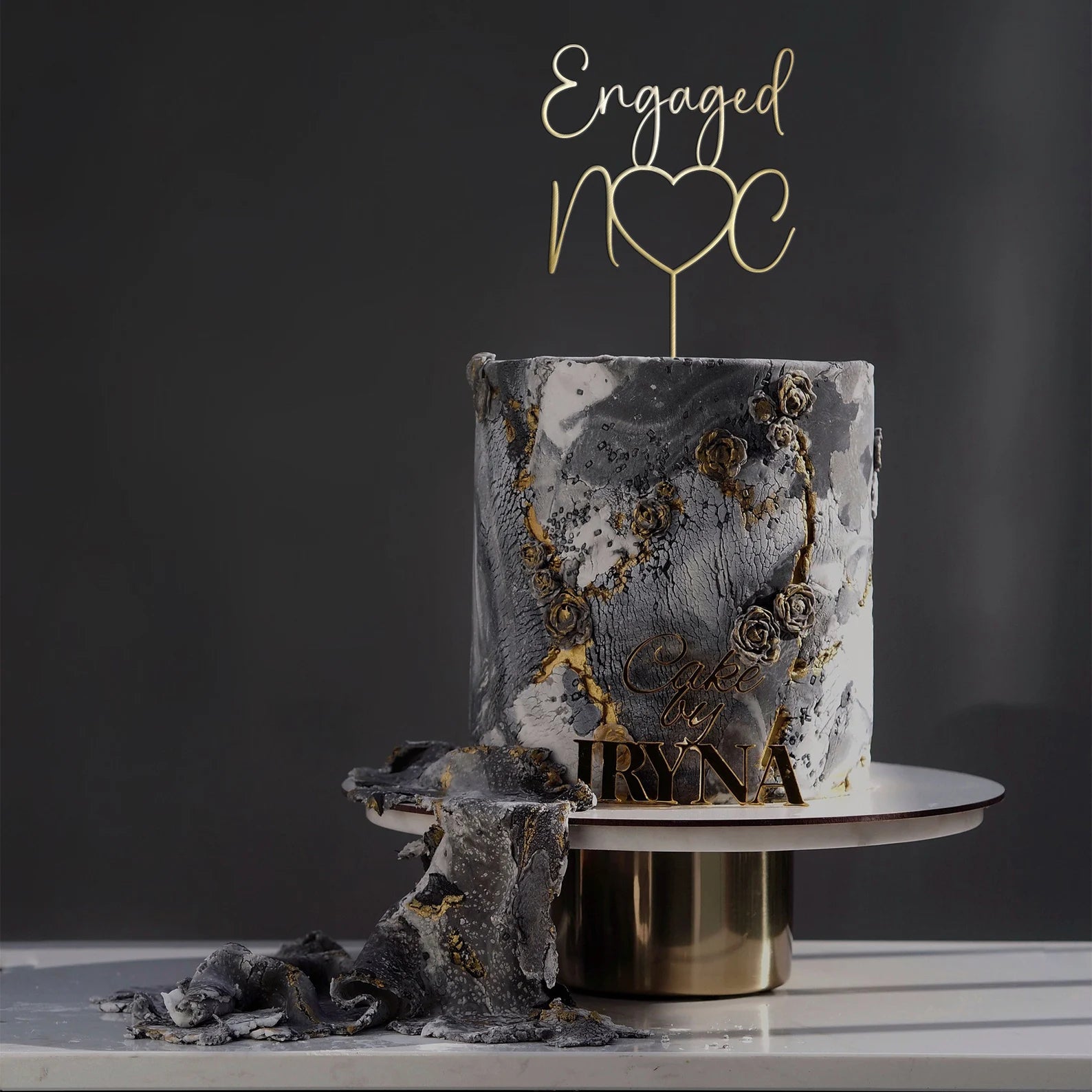 A two-tiered cake featuring a marbled gray and white design with gold accents and floral decorations. Topping the cake is a personalized acrylic cake topper that reads "Engaged" with a heart, displayed on a circular cake stand with a gold base. A piece of cake has been cut, showing the inside. The background is dark, enhancing the elegant presentation of the cake.