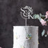A close-up of a white, textured wedding cake featuring pastel-colored flower decorations. Atop the cake is a custom acrylic cake topper displayed in silver, featuring the initials "S&K" and the date "24.04.2024," accompanied by a delicate leaf design. The background is softly blurred, enhancing the focus on the cake and topper.