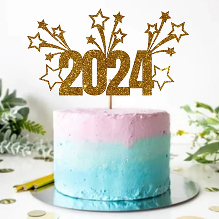 A colorful cake with a gradient design features a light pink top layer and a light blue bottom layer. A gold glittery cake topper displays the year "2024" along with decorative stars and bursts. The cake is placed on a round silver cake board, surrounded by green foliage and scattered gold confetti on the table.