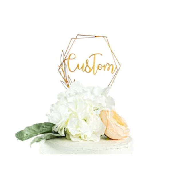 A geometric acrylic cake topper featuring the word "Custom" in a stylish golden font, situated above a decorated cake. The cake is adorned with white flowers and a light peach-colored rose, along with green leaves. The topper has a hexagonal shape.