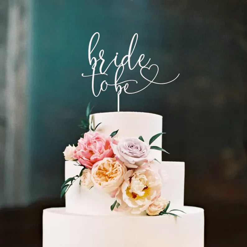 A three-tiered white cake adorned with a floral arrangement featuring pink and purple roses, as well as peonies. At the top, a scripted acrylic cake topper reads "bride to be," with a decorative heart. The background is softly blurred, enhancing the cake&