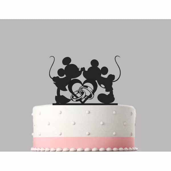 A white cake topped with a black acrylic cake topper featuring two cartoon mouse silhouettes, one holding hands with the other. At the center, there is a intertwined heart design. The cake has a pink ribbon around its base and is decorated with white fondant dots. The background is gray.