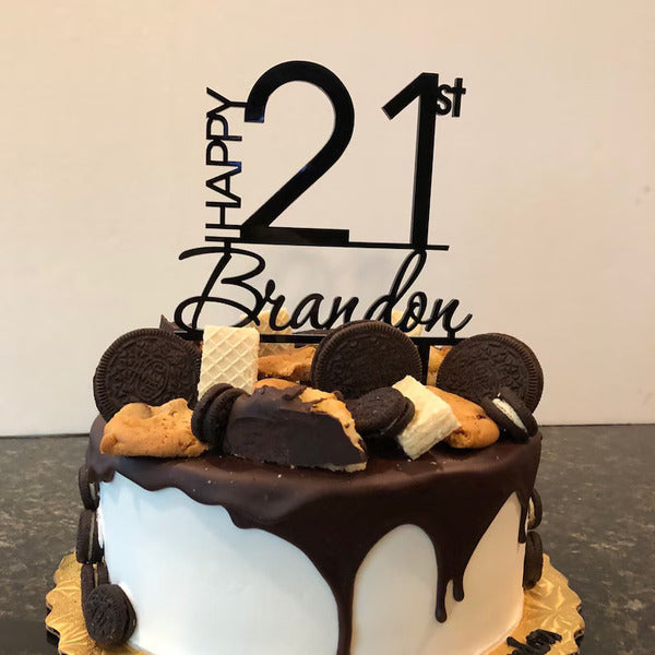 A round birthday cake topped with various types of cookies and chocolate decorations. Atop the cake is a black cake topper that reads "HAPPY 21st Brandon." The cake has a white frosting base with a dripping chocolate ganache effect. The decorations include Oreos, chocolate chip cookies, and waffle cones, all arranged artistically on the surface. The cake sits on a golden-colored cake board, with a neutral background.