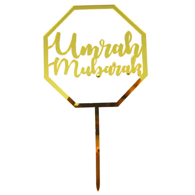 A gold acrylic cake topper in the shape of an octagon with the words "Umrah Mubarak" written in elegant, cursive font. It features a pointed stick at the bottom for easy placement on cakes or desserts.