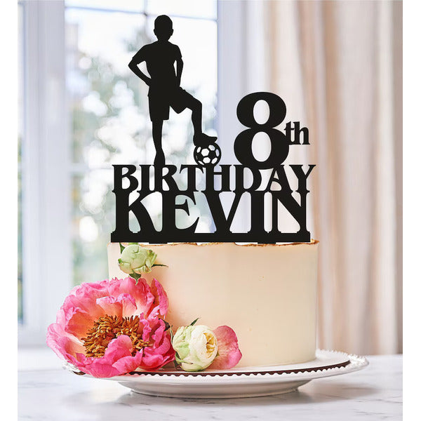 A decorated cake featuring a cream-colored frosting is topped with a black personalized soccer player cake topper. The topper includes the text "8th BIRTHDAY KEVIN," and a silhouette of a soccer player in a standing pose, one foot on a soccer ball. Surrounding the base of the topper are vibrant flowers, including pink peonies and green accents. The cake is presented on a decorative white plate. The background suggests a bright and cheerful indoor setting.