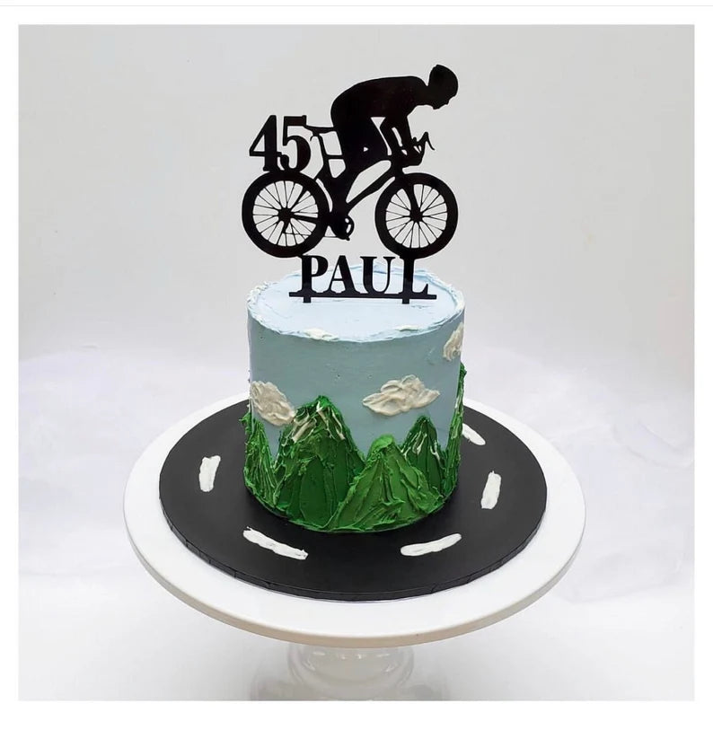 A decorated cake featuring a blue frosting top with mountain designs and clouds, a green grass-like texture at the base, and a black silhouette of a cyclist riding a bicycle on top, along with the number "45" and the name "PAUL" in black lettering. The cake sits on a round black base with white road markings.