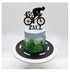 A decorated cake featuring a blue frosting top with mountain designs and clouds, a green grass-like texture at the base, and a black silhouette of a cyclist riding a bicycle on top, along with the number "45" and the name "PAUL" in black lettering. The cake sits on a round black base with white road markings.
