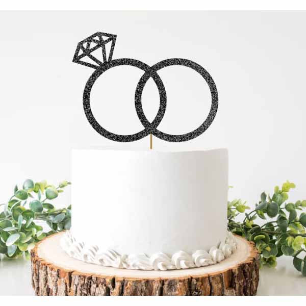 A round white cake is centered on a rustic wooden cake stand. Topping the cake is a black glittery cake topper featuring two interlocking rings, with one ring adorned by a diamond-shaped design. The decorated cake is surrounded by green foliage, adding a touch of color to the presentation.