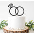 A round white cake is centered on a rustic wooden cake stand. Topping the cake is a black glittery cake topper featuring two interlocking rings, with one ring adorned by a diamond-shaped design. The decorated cake is surrounded by green foliage, adding a touch of color to the presentation.