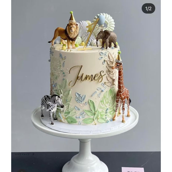 A decorated cake featuring a fun, jungle-themed design. The cake is white with green leaf accents, a personalized name "James" in gold lettering, and is topped with various animal figurines, including a lion, a giraffe, an elephant, and a zebra. Colorful decorative elements like party hats and a cake topper complete the festive look. The cake is displayed on a white cake stand.