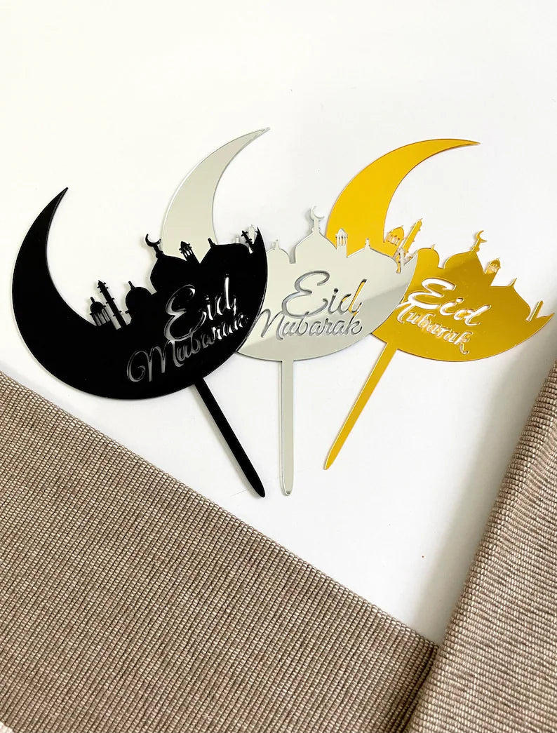 The image features three acrylic cake toppers designed for Eid celebrations. Each topper is shaped like a crescent moon with a silhouette of a mosque and features the text "Eid Mubarak." The toppers are in three distinct colors: black, silver, and gold. They are displayed on a neutral-colored textured surface, with one corner slightly covered by a fabric.