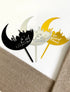The image features three acrylic cake toppers designed for Eid celebrations. Each topper is shaped like a crescent moon with a silhouette of a mosque and features the text "Eid Mubarak." The toppers are in three distinct colors: black, silver, and gold. They are displayed on a neutral-colored textured surface, with one corner slightly covered by a fabric.