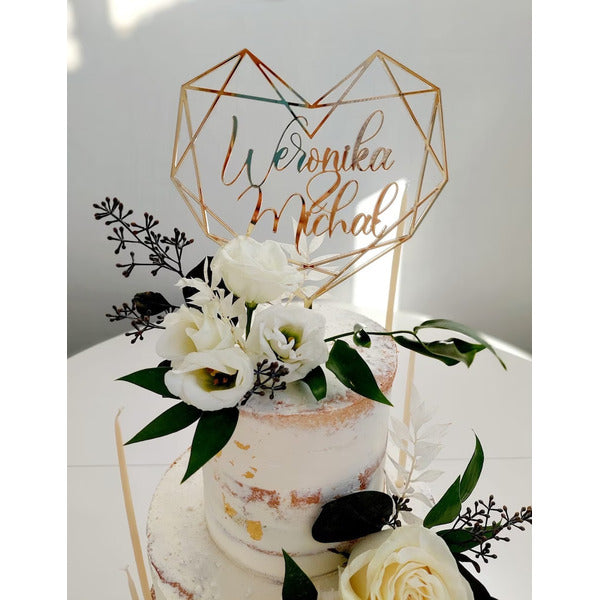 The image depicts a two-tier wedding cake topped with a personalized cake topper. The topper features a geometric heart design made of gold, with the names "Weronika" and "Michal" elegantly displayed in a cursive font. Surrounding the base of the topper are decorative white roses and greenery that complement the cake&