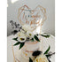 The image depicts a two-tier wedding cake topped with a personalized cake topper. The topper features a geometric heart design made of gold, with the names "Weronika" and "Michal" elegantly displayed in a cursive font. Surrounding the base of the topper are decorative white roses and greenery that complement the cake&