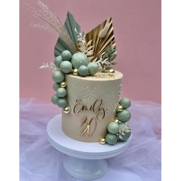 A round, ombre cake decorated with a textured light beige finish. The cake is adorned with green and gold spherical decorations and a mix of foliage, including a large green leaf and dried grasses. A personalized topper featuring the name "Emily" in an elegant script and the number "20" in a golden finish is prominently displayed on the front. The cake is placed on a white cake stand against a soft pink background.