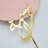 A gold acrylic cake topper featuring an elegant design, resting on a light pink plate. The topper has a stylized script that reads "Happy Birthday" in Arabic. The cake topper has a pointed stick for easy placement on cakes.