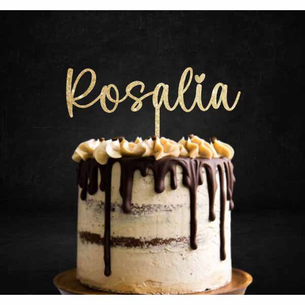 A decorated cake with a cream-colored frosting and chocolate ganache dripping down the sides. The cake is topped with a swirl of cream and banana slices. Above the cake, there is a sparkling gold glitter cake topper that reads "Rosalia" in a cursive font. The background is dark, enhancing the visual appeal of the cake.