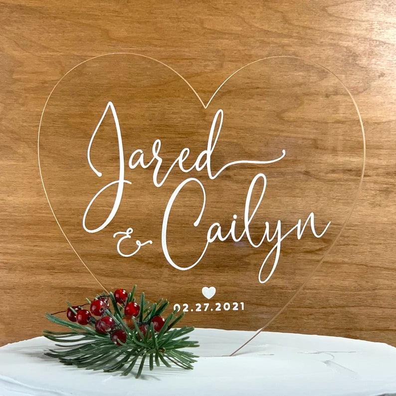 The image features an acrylic wedding cake topper in the shape of a heart. The topper displays the names "Jared" and "Cailyn" in stylish, elegant white script. Below the names, the date "02.27.2021" is included, accompanied by a small heart graphic. The topper is placed on a white cake, with a decorative arrangement of greenery and red berries underneath it. The background is a wooden surface, enhancing the overall rustic and romantic appearance.
