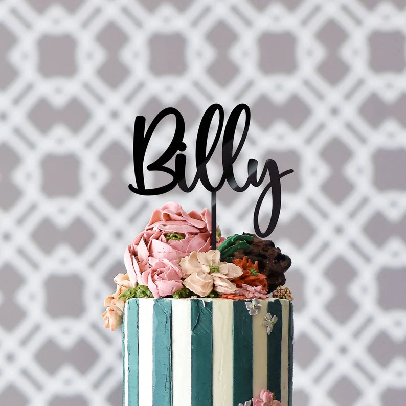 A personalized acrylic cake topper featuring the name "Billy" is placed atop a beautifully decorated cake. The cake has alternating dark blue and white vertical stripes and is adorned with colorful, delicate floral decorations in shades of pink, cream, and green. The background consists of a subtle, patterned design that adds an elegant touch to the overall presentation.