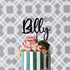 A personalized acrylic cake topper featuring the name "Billy" is placed atop a beautifully decorated cake. The cake has alternating dark blue and white vertical stripes and is adorned with colorful, delicate floral decorations in shades of pink, cream, and green. The background consists of a subtle, patterned design that adds an elegant touch to the overall presentation.