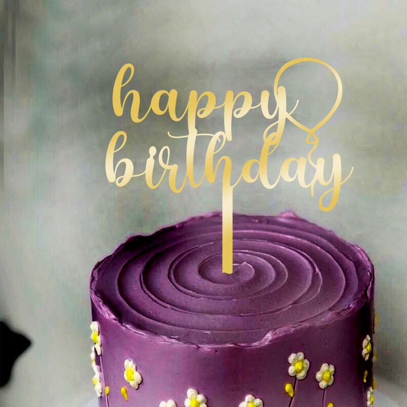 An acrylic cake topper featuring the words "happy birthday" in elegant, gold script is placed atop a purple frosted cake. The cake has a spiraled icing design and is decorated with small white flowers. The background is a soft, blurred color.