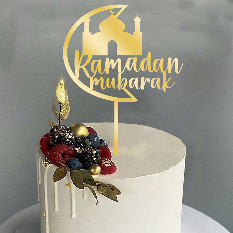 A round acrylic cake topper featuring the words "Ramadan Mubarak" in elegant script, accompanied by a silhouette of a mosque. The topper is gold-colored and is placed on top of a white frosted cake, decorated with a colorful assortment of berries, including raspberries and blueberries, along with golden embellishments and green leaves. The cake has a smooth finish and a dripping icing effect on the sides. The background is softly blurred.