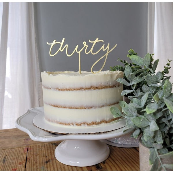 A three-layer cake with a creamy white frosting and golden-brown layers is displayed on a white cake stand. Atop the cake is a decorative acrylic topper that reads "thirty" in an elegant, cursive font. To the right of the cake is a small green plant, enhancing the presentation. The backdrop features soft, neutral-toned curtains, adding to the overall aesthetic.