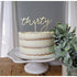 A three-layer cake with a creamy white frosting and golden-brown layers is displayed on a white cake stand. Atop the cake is a decorative acrylic topper that reads "thirty" in an elegant, cursive font. To the right of the cake is a small green plant, enhancing the presentation. The backdrop features soft, neutral-toned curtains, adding to the overall aesthetic.
