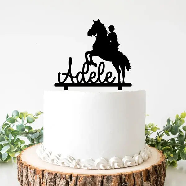 A personalized cake topper featuring a silhouette of a horse and rider is placed on top of a round white cake. The rider is depicted in a mounted position, and below the silhouette, the name "Adele" is written in a flowing script. The cake is set on a wooden slice and surrounded by greenery.