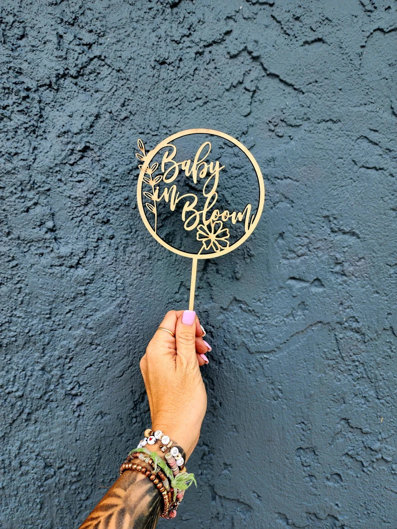 A hand is holding an acrylic cake topper that features the words "Baby in Bloom" inside a circular design. The topper includes decorative elements, such as flowers and leaves. The hand is adorned with multiple colorful bracelets, and the background is a textured dark blue wall.