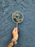 A hand is holding an acrylic cake topper that features the words "Baby in Bloom" inside a circular design. The topper includes decorative elements, such as flowers and leaves. The hand is adorned with multiple colorful bracelets, and the background is a textured dark blue wall.