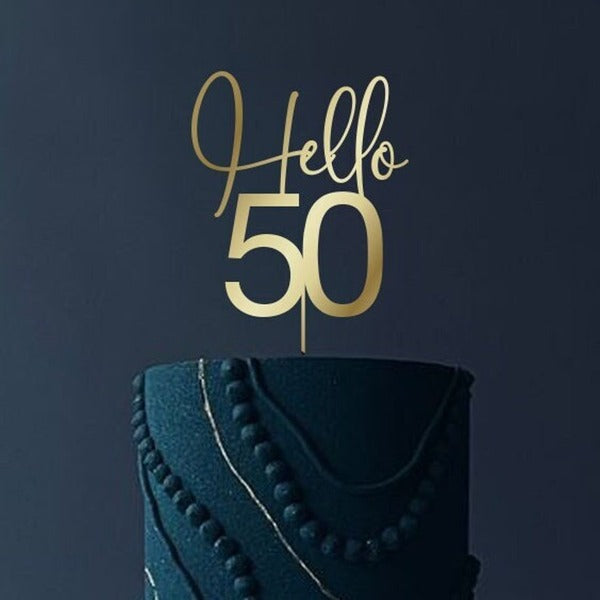 A decorative cake topper made of acrylic, featuring the words "Hello" in a cursive font and the number "50" in a bold font. The topper is placed on a dark, textured birthday cake that is decorated with beaded and swirling patterns in a matching color. The background is dark, emphasizing the cake and the topper.