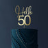 A decorative cake topper made of acrylic, featuring the words "Hello" in a cursive font and the number "50" in a bold font. The topper is placed on a dark, textured birthday cake that is decorated with beaded and swirling patterns in a matching color. The background is dark, emphasizing the cake and the topper.