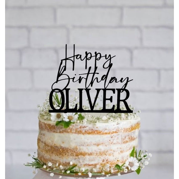 A round, multi-layered cake is decorated with a black acrylic cake topper that reads "Happy Birthday OLIVER" in a combination of script and bold font. The cake has a rustic appearance, with visible layers and light frosting. It is adorned with small white flowers and greenery around the base. The background features a simple, light-colored brick wall, enhancing the festive feel of the scene.