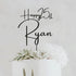 A personalized acrylic cake topper is displayed, featuring the text "Happy 25th Ryan" in an elegant, cursive font. The topper is positioned on a white cake adorned with succulent decorations and white flowers. The background is light and neutral, emphasizing the topper and the cake.