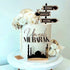 The image features a decorative cake with a cream-colored exterior. At the front, the words "Umrah Mubarak" are beautifully crafted in an elegant font. The cake is adorned with white and pale floral arrangements along with golden accents. Topping the cake, there is a signpost with three arrows indicating the directions to "Jeddah," "Makkah," and "Madina." Additionally, a silhouette of a mosque is depicted at the base of the cake, enhancing its festive theme. The cake is displayed on a white round stand.
