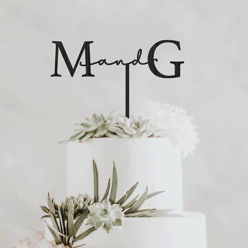 A three-tier white wedding cake decorated with greenery and succulents. The cake features a black personalized cake topper displaying the initials "M" and "G" connected by the word "and" in a cursive font. The background is soft and neutral, enhancing the elegance of the cake design.