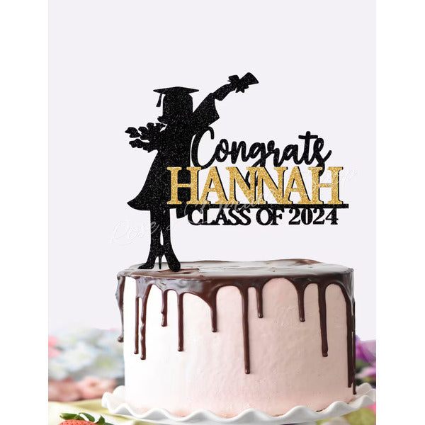The image features a cake decorated with a pink frosting and chocolate ganache drizzled on top. A decorative cake topper is prominently displayed on the cake, which includes the words "Congrate HANNAH" in a mix of black and glittery gold letters, along with "CLASS OF 2024." The topper features a silhouette of a graduate wearing a graduation cap and gown, holding a bouquet of flowers and a diploma. The background includes a blurred setting, suggesting a celebratory atmosphere.